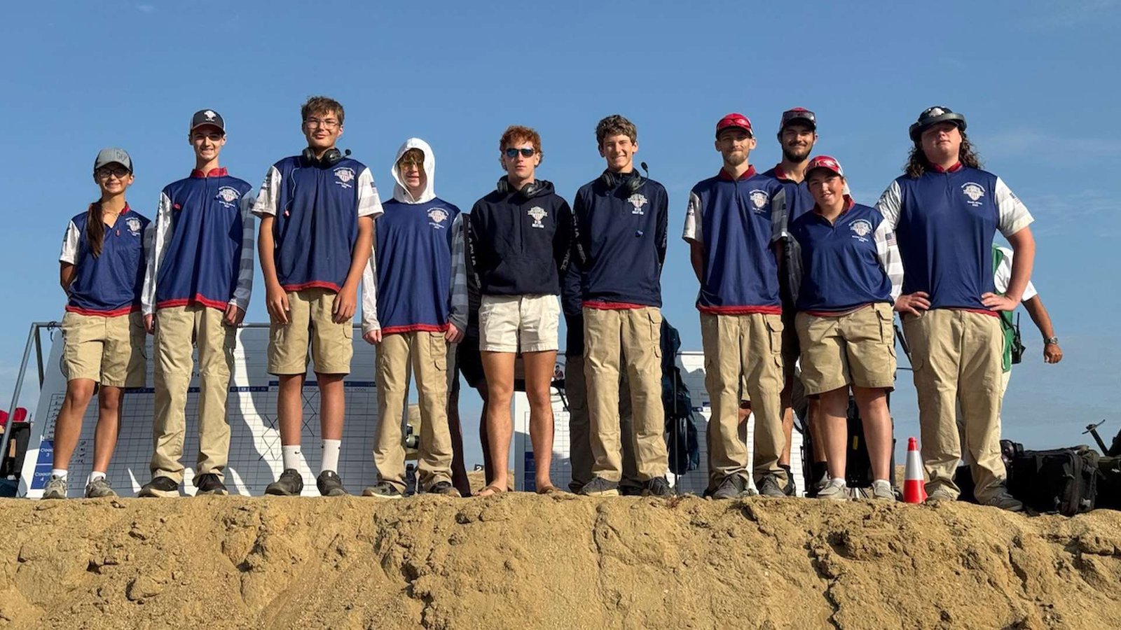 Ten young American shooters in team jerseys standing along a berm on the range at Bisley under a clear sky